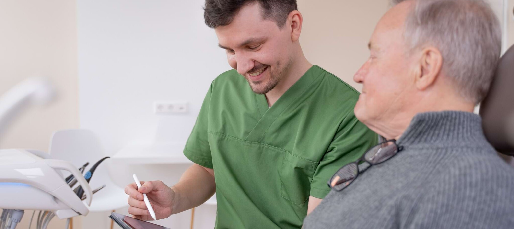 Fotoaufnahme: Christopher Schwenn zeigt einem Patienten im Behandlungsstuhl etwas auf einem Tablet