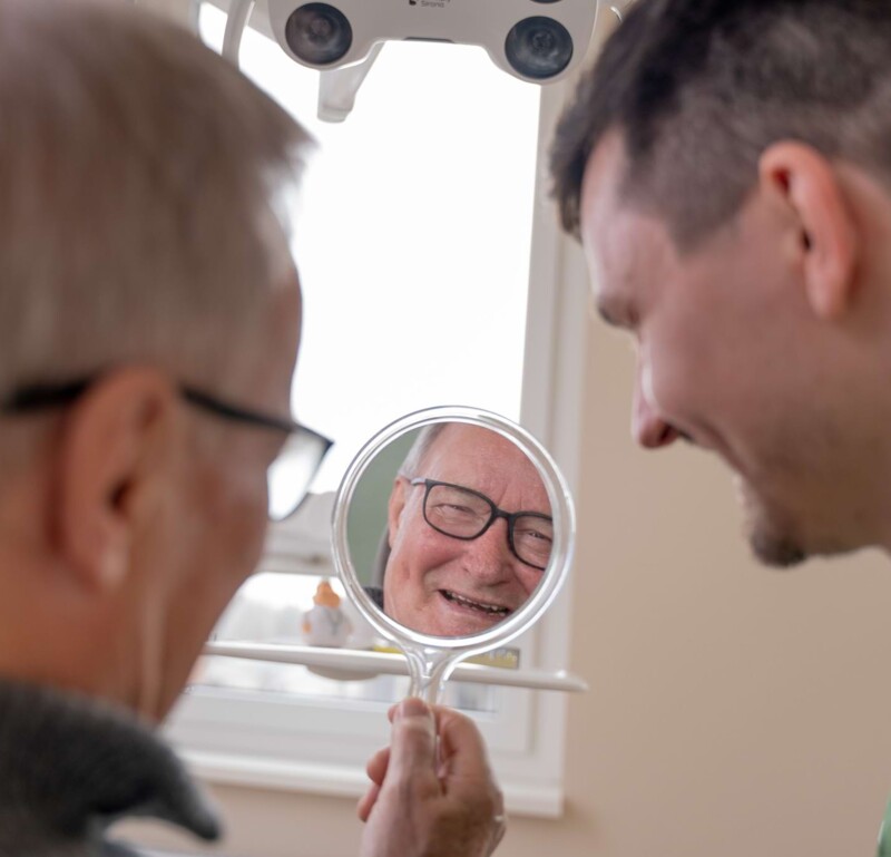 Fotoaufnahme: Christopher Schwenn und Patient lachen gemeinsam, der Patient schaut in einen Handspiegel