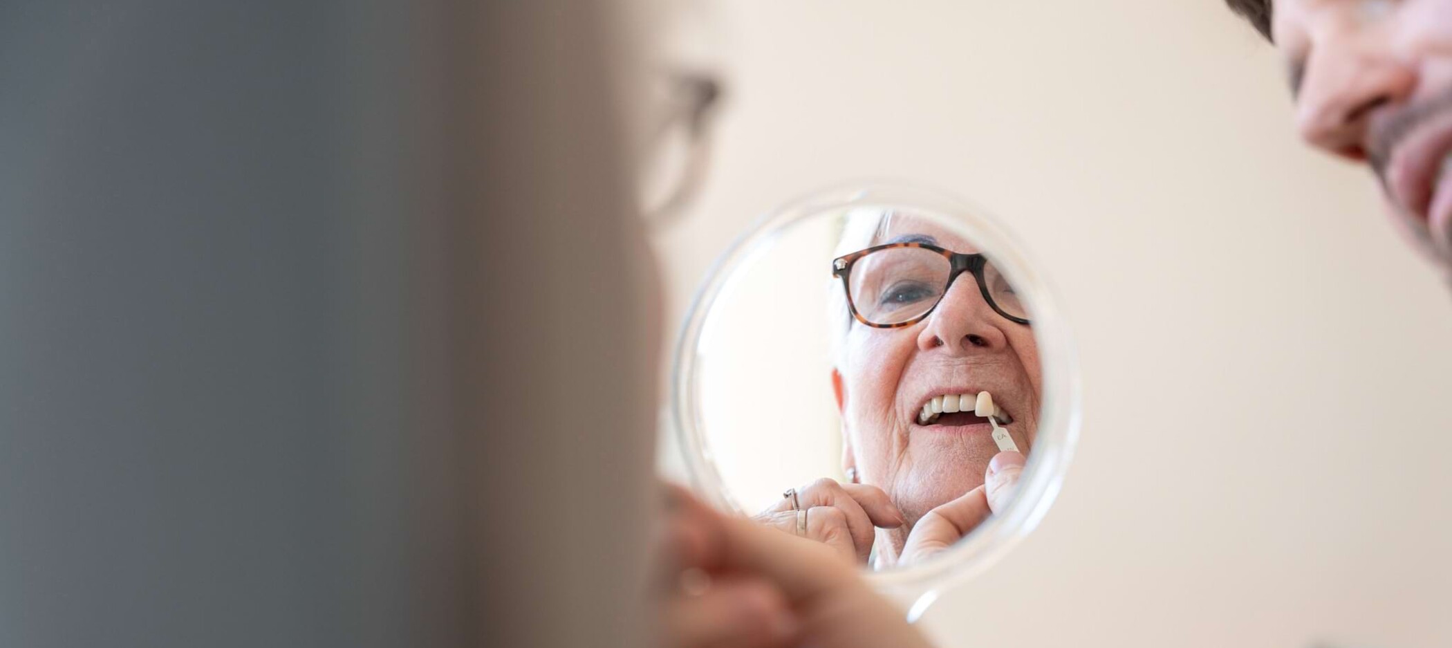 Fotoaufnahme: Patientin, der ein Zahnimplantat vor die Zähne gehalten wird, hält einen Handspiegel, Fokus auf den Spiegel