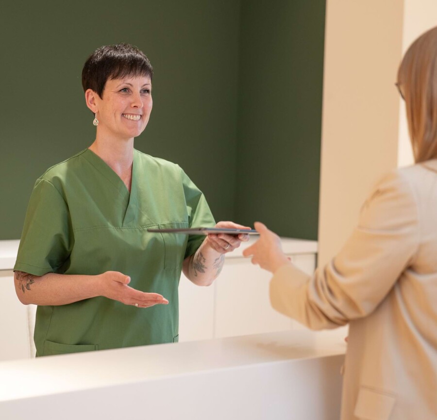 Fotoaufnahme: Mitarbeiterin übergibt einer Patientin am Empfang Unterlagen