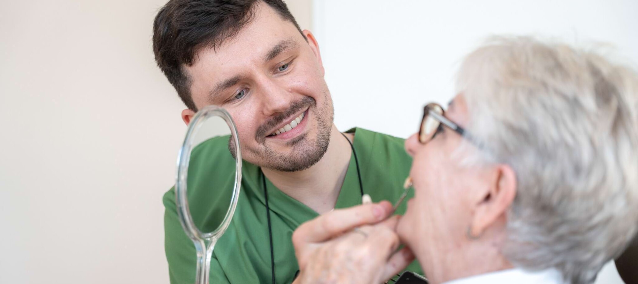 Fotoaufnahme: Christopher Schwenn hält ein Zahnimplantat testweise vor eine Patientin, die einen Handspiegel hält