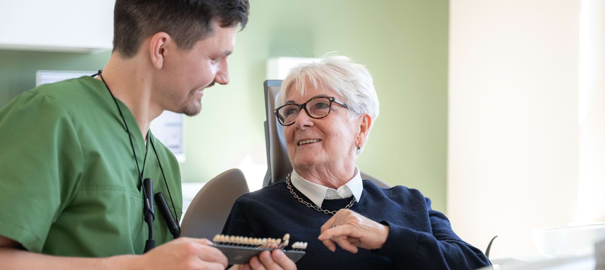 Fotoaufnahme: Christopher Schwenn hält eine Zahnfarbskala und berät eine Patientin im Behandlungsstuhl